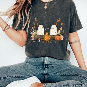 Puede incluir: Camiseta gris oscuro con una ilustraci&oacute;n de dos fantasmas blancos asando malvaviscos sobre una fogata. El dise&ntilde;o incluye un tronco, una calabaza y hojas de oto&ntilde;o, ideal para Halloween o el oto&ntilde;o.