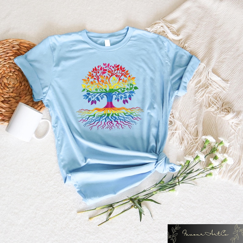 May include: Light blue t-shirt featuring a colorful tree design. The tree's leaves, trunk, and roots are rendered in a rainbow of colors. The shirt is made of soft material and is suitable for casual wear.