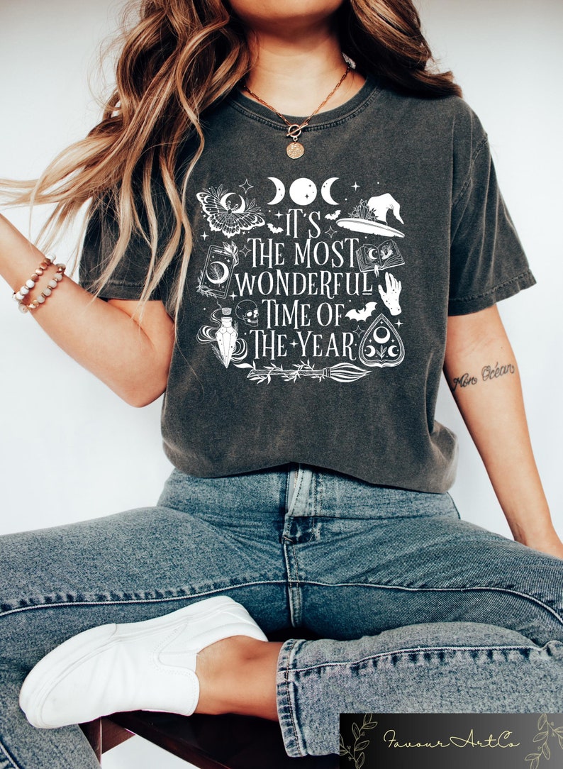 May include: A dark gray t-shirt with a white graphic design featuring a witch's hat, a crescent moon, a potion bottle, a skull, a bat, and the text "It's the most wonderful time of the year".
