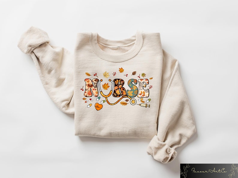 Pode incluir: Uma camisa de moletom bege com a palavra "NURSE" impressa em letras coloridas com um design de outono. As letras s&atilde;o decoradas com folhas, flores e s&iacute;mbolos m&eacute;dicos como um estetosc&oacute;pio e uma seringa.