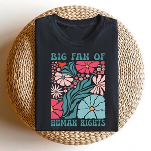May include: Black t-shirt with a floral design and the text "BIG FAN OF HUMAN RIGHTS" in teal. The floral design includes pink, teal, and white flowers and green leaves. The shirt is displayed on a woven, circular surface.