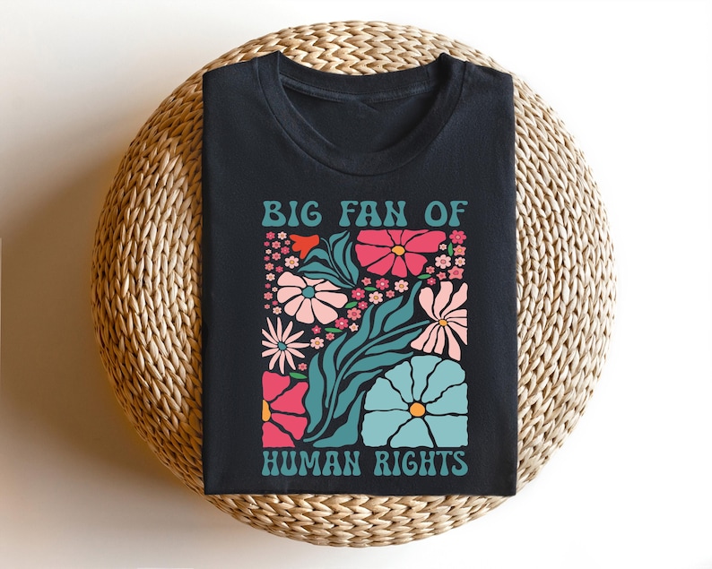 May include: Black t-shirt with a floral design and the text "BIG FAN OF HUMAN RIGHTS" in teal. The floral design includes pink, teal, and white flowers and green leaves. The shirt is displayed on a woven, circular surface.