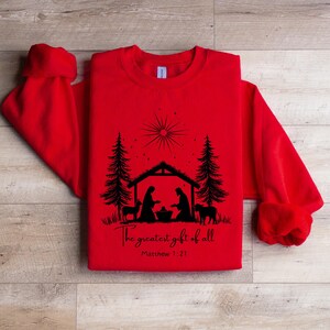May include: Red sweatshirt with a black graphic of a nativity scene, including silhouettes of Mary, Joseph, baby Jesus, animals, and trees. The text "The greatest gift of all" and Matthew 1:21 are printed below the image.