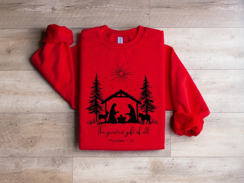 May include: Red sweatshirt with a black graphic of a nativity scene, including silhouettes of Mary, Joseph, baby Jesus, animals, and trees. The text "The greatest gift of all" and Matthew 1:21 are printed below the image.