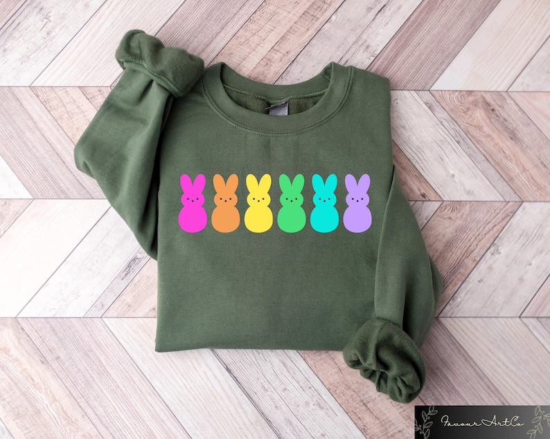 K&ouml;nnte beinhalten: Ein gr&uuml;ner Pullover mit einer Reihe von sechs bunten, kaninchenf&ouml;rmigen Marshmallow-Peeps. Die Peeps sind rosa, orange, gelb, gr&uuml;n, blau und lila.