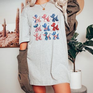 K&ouml;nnte beinhalten: Hellgraues T-Shirt mit einem Raster von neun Ballonhund-Illustrationen. Die Hunde sind in Rot, Wei&szlig; und Blau gehalten, mit Sternen- und Streifenmustern. Das Shirt hat einen l&auml;ssigen, &uuml;bergro&szlig;en Stil.