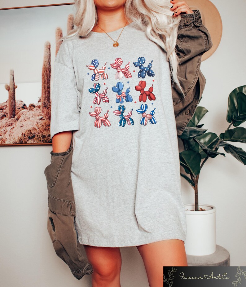 K&ouml;nnte beinhalten: Hellgraues T-Shirt mit einem Raster von neun Ballonhund-Illustrationen. Die Hunde sind in Rot, Wei&szlig; und Blau gehalten, mit Sternen- und Streifenmustern. Das Shirt hat einen l&auml;ssigen, &uuml;bergro&szlig;en Stil.