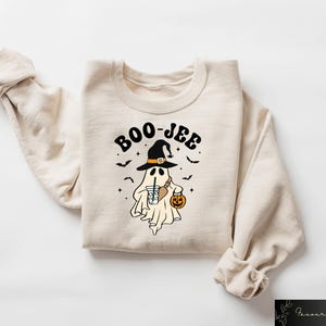 Może przedstawiać: Kremowa bluza z nadrukiem przedstawiającym ducha w czarodziejskim kapeluszu, trzymającego dynię i nap&oacute;j. Nad duchem widnieje napis "BOO-JEE".