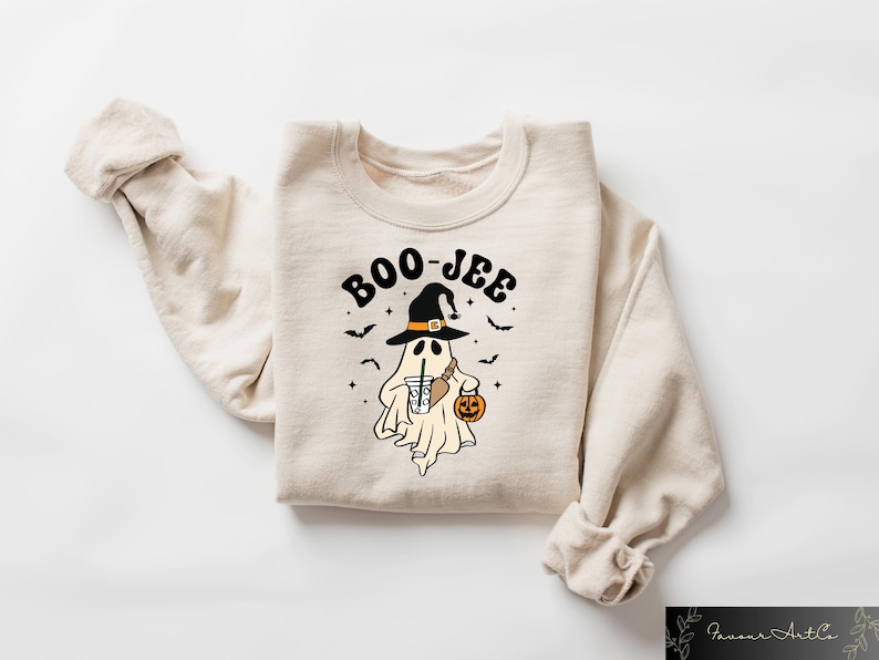 Może przedstawiać: Kremowa bluza z nadrukiem przedstawiającym ducha w czarodziejskim kapeluszu, trzymającego dynię i nap&oacute;j. Nad duchem widnieje napis "BOO-JEE".