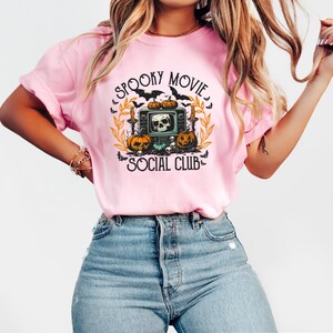 Puede incluir: Camiseta rosa claro con un dise&ntilde;o de Halloween. El dise&ntilde;o presenta una calavera dentro de un televisor retro, calabazas, murci&eacute;lagos y el texto "Spooky Movie Social Club."