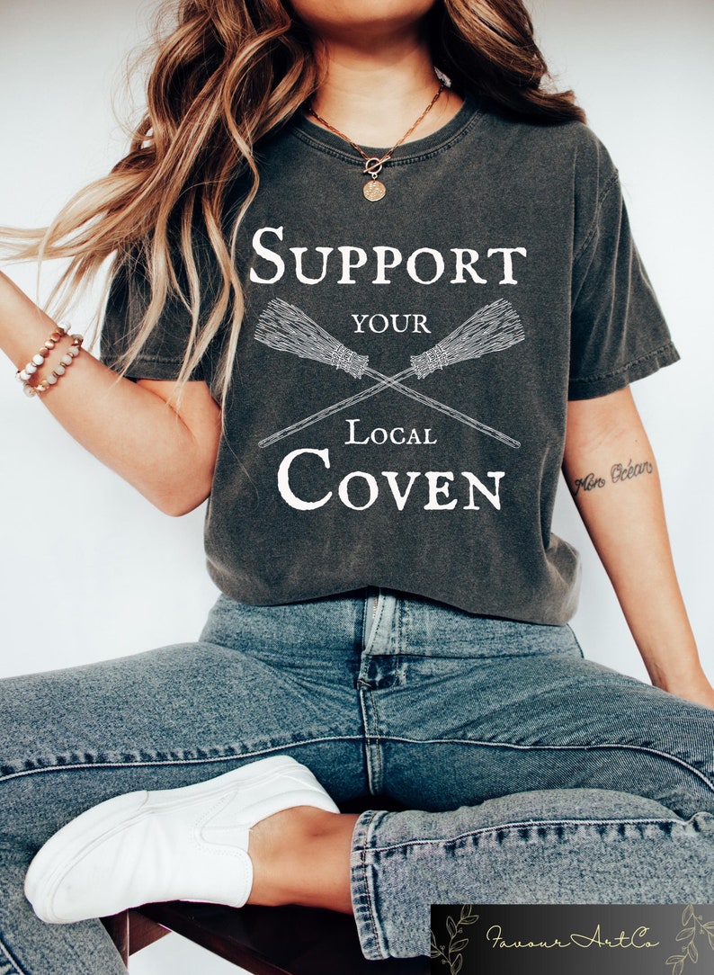 May include: A dark gray t-shirt with a white graphic that says "Support Your Local Coven" with two crossed broomsticks.