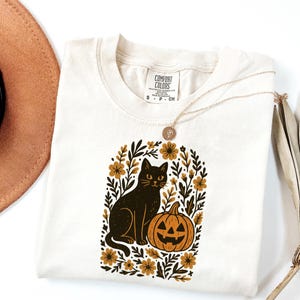 May include: Cream-colored t-shirt featuring a black cat and jack-o'-lantern graphic within a floral arch. The design includes gold and brown accents. The shirt has a Comfort Colors tag and a gold necklace.