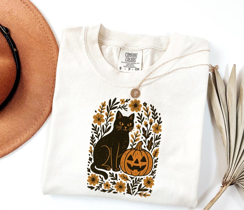 May include: Cream-colored t-shirt featuring a black cat and jack-o'-lantern graphic within a floral arch. The design includes gold and brown accents. The shirt has a Comfort Colors tag and a gold necklace.