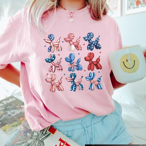 K&ouml;nnte beinhalten: Ein hellrosa T-Shirt mit einem sich wiederholenden Muster aus Ballonhund-Designs in Rot, Wei&szlig; und Blau, mit Sternakzenten. Das Shirt ist ein l&auml;ssiger Stil, geeignet f&uuml;r den Alltag.