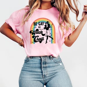 Pode incluir: Camiseta rosa claro com o texto "BE GAY DO Witch Craft" acima de um desenho de uma bruxa em frente a um arco-&iacute;ris. A camiseta &eacute; rosa claro e tem mangas curtas.