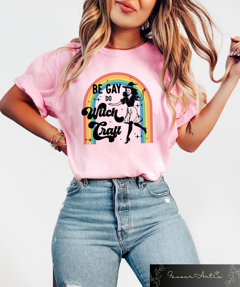 Pode incluir: Camiseta rosa claro com o texto "BE GAY DO Witch Craft" acima de um desenho de uma bruxa em frente a um arco-&iacute;ris. A camiseta &eacute; rosa claro e tem mangas curtas.