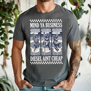 May include: Gray t-shirt with a graphic featuring three raccoons on baseball poles, with the text "MIND YA BUSINESS" above and "DIESEL AINT CHEAP" below in a white font. The shirt has a crew neck and short sleeves.