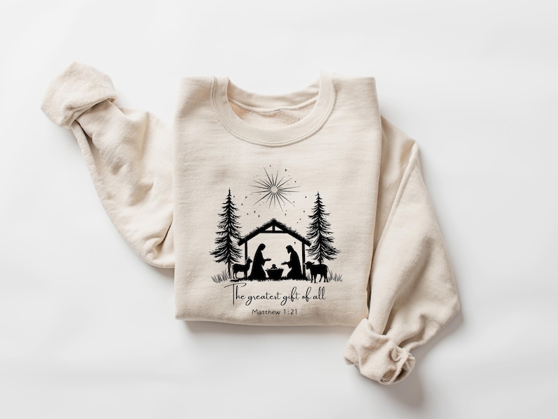 May include: Cream-colored sweatshirt with a black graphic of a nativity scene, including a star, trees, and animals. The text "The greatest gift of all" and Matthew 1:21 are printed below the image. A cozy, long-sleeved top.