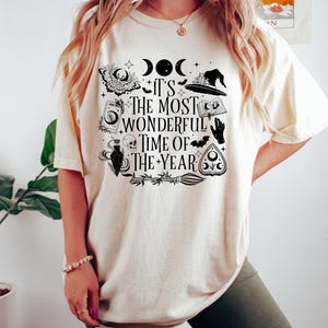 May include: A cream colored t-shirt with a black graphic print of a witch's hat, a crescent moon, a skull, a potion bottle, a bat, a hand, a book, a broom, and a pentagram. The text on the shirt reads "It's the most wonderful time of the year."