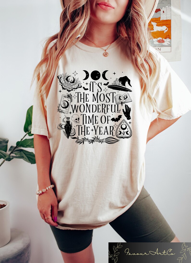 May include: A cream colored t-shirt with a black graphic print of a witch's hat, a crescent moon, a skull, a potion bottle, a bat, a hand, a book, a broom, and a pentagram. The text on the shirt reads "It's the most wonderful time of the year."