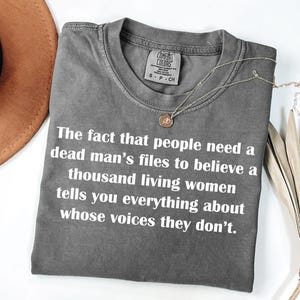 May include: A gray Comfort Colors t-shirt with white text: "The fact that people need a dead man's files to believe a thousand living women tells you everything about whose voices they don't." A gold necklace is draped across the shirt.