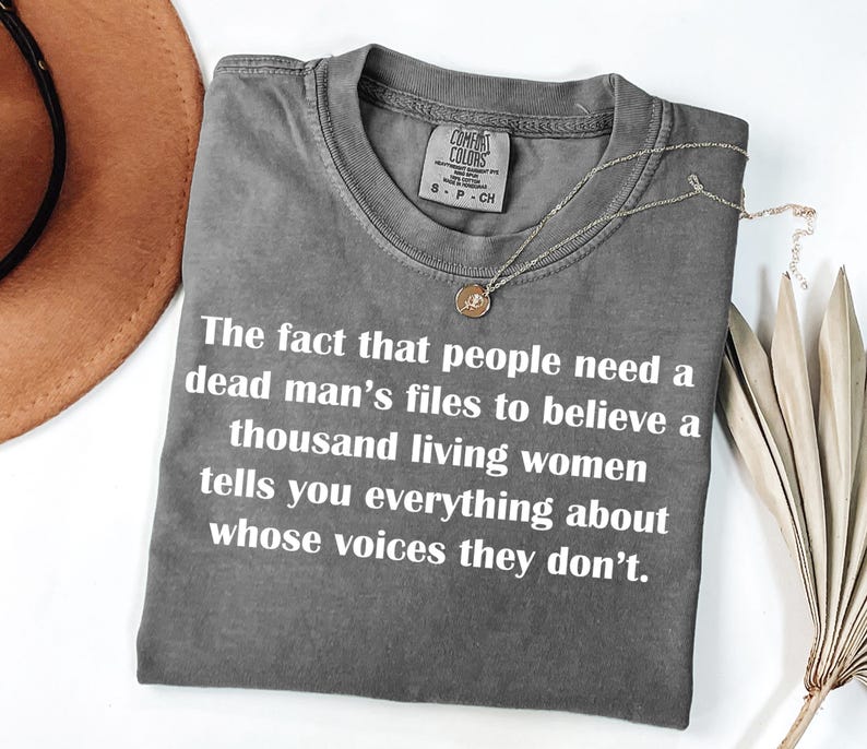 May include: A gray Comfort Colors t-shirt with white text: "The fact that people need a dead man's files to believe a thousand living women tells you everything about whose voices they don't." A gold necklace is draped across the shirt.