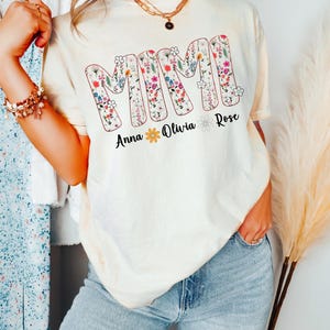 Pode incluir: Camiseta creme com a palavra "MIMI" em letras grandes e floridas. Abaixo da palavra est&atilde;o os nomes "Anna", "Olivia" e "Rose". A camiseta &eacute; usada com jeans azuis.