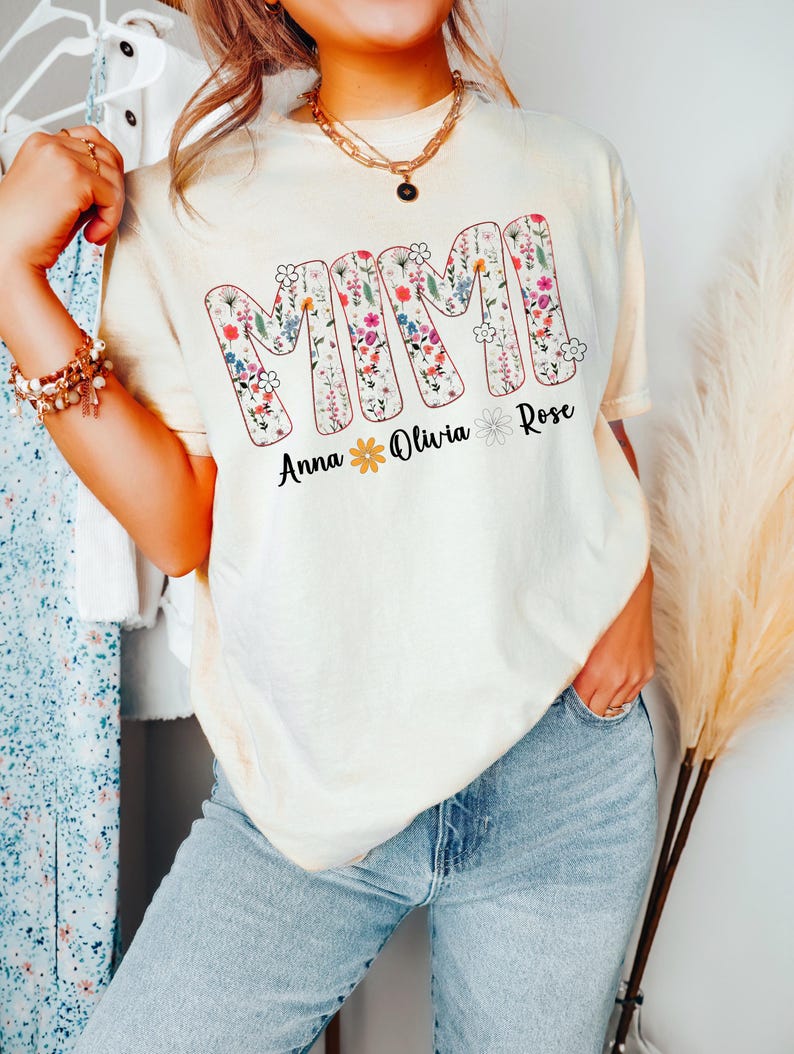 Pode incluir: Camiseta creme com a palavra "MIMI" em letras grandes e floridas. Abaixo da palavra est&atilde;o os nomes "Anna", "Olivia" e "Rose". A camiseta &eacute; usada com jeans azuis.