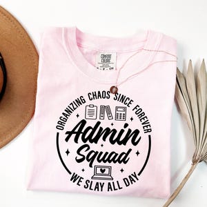 May include: A pale pink t-shirt featuring the text "Organizing Chaos Since Forever" and "Admin Squad We Slay All Day" in a black circular design. The design includes a clipboard, files, and a calculator. A gold necklace is draped over the shirt.