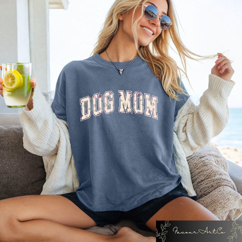 May include: A woman wearing a blue t-shirt with the text "DOG MOM" in white letters with a floral pattern. The shirt has a relaxed fit and is perfect for casual wear.