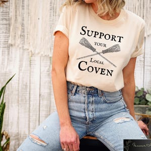 May include: A beige t-shirt with the text "Support Your Local Coven" and a graphic of two crossed brooms.