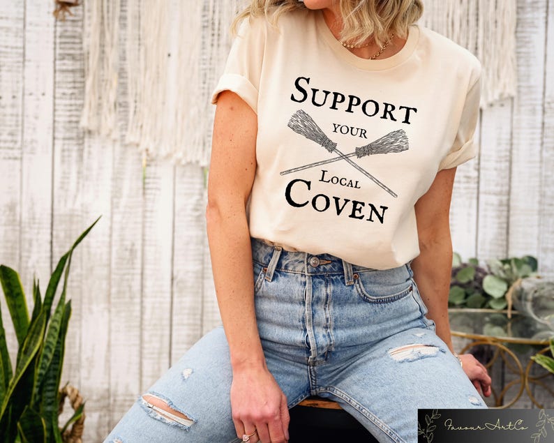 May include: A beige t-shirt with the text "Support Your Local Coven" and a graphic of two crossed brooms.