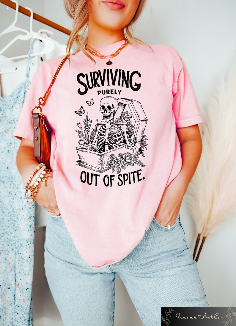 May include: Pink t-shirt with a black and white graphic of a skeleton sitting in a coffin with flowers and butterflies. The text on the shirt reads "SURVIVING PURELY OUT OF SPITE."