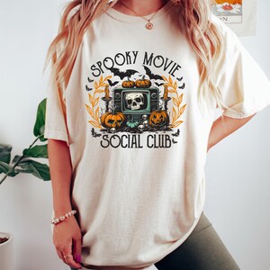 Puede incluir: Camiseta color crema con un dise&ntilde;o tem&aacute;tico de Halloween. El dise&ntilde;o presenta un televisor vintage con una calavera, calabazas, murci&eacute;lagos y las palabras "Spooky Movie Social Club."