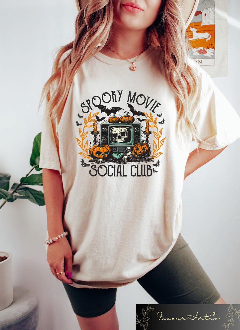Puede incluir: Camiseta color crema con un dise&ntilde;o tem&aacute;tico de Halloween. El dise&ntilde;o presenta un televisor vintage con una calavera, calabazas, murci&eacute;lagos y las palabras "Spooky Movie Social Club."
