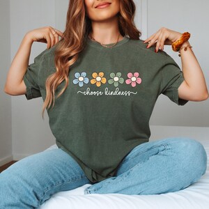 Puede incluir: Camiseta verde oliva con un dise&ntilde;o floral con flores azules, amarillas, verdes y rosas. Las palabras "choose kindness" est&aacute;n escritas en cursiva debajo de las flores. La camiseta se combina con unos vaqueros azules.