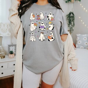 May include: A gray t-shirt featuring a whimsical design of cartoon ghosts reading books. The ghosts are white with black eyes and are holding books of various colors. The design is surrounded by small star shapes.