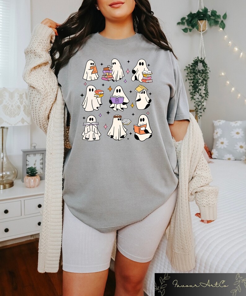 May include: A gray t-shirt featuring a whimsical design of cartoon ghosts reading books. The ghosts are white with black eyes and are holding books of various colors. The design is surrounded by small star shapes.
