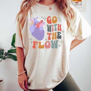 May include: A white t-shirt with a colorful graphic of a heart and the text "GO WITH THE FLOW" in a retro font.