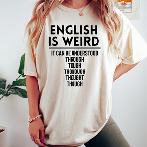 May include: A white t-shirt with the text "ENGLISH IS WEIRD" and the phrase "IT CAN BE UNDERSTOOD THROUGH TOUGH THOROUGH THOUGHT THOUGH" printed in black.