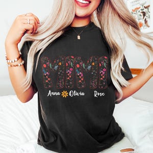 Pode incluir: Camiseta preta com a palavra "MIMI" em um design floral. Abaixo, os nomes "Anna", "Olivia" e "Rose" est&atilde;o escritos. A camiseta &eacute; escura e est&aacute; sendo usada por uma pessoa.
