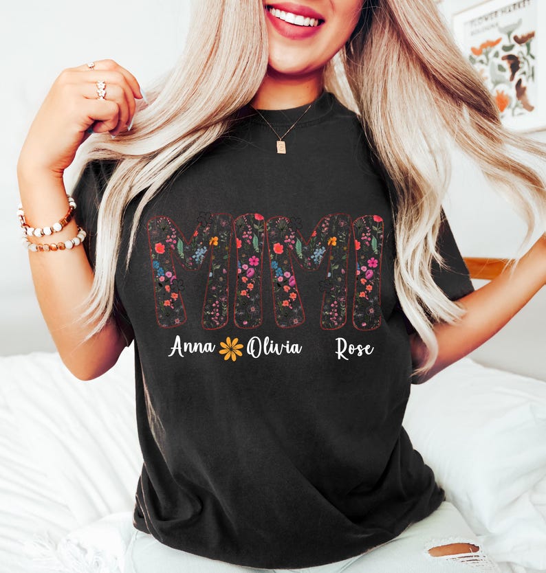 Pode incluir: Camiseta preta com a palavra "MIMI" em um design floral. Abaixo, os nomes "Anna", "Olivia" e "Rose" est&atilde;o escritos. A camiseta &eacute; escura e est&aacute; sendo usada por uma pessoa.