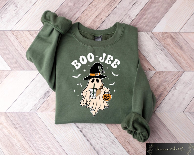Może przedstawiać: Zielona bluza z nadrukiem białego ducha w czarodziejskim kapeluszu, trzymającego dynię i kubek kawy. Na bluzie widnieje napis "BOO-JEE".