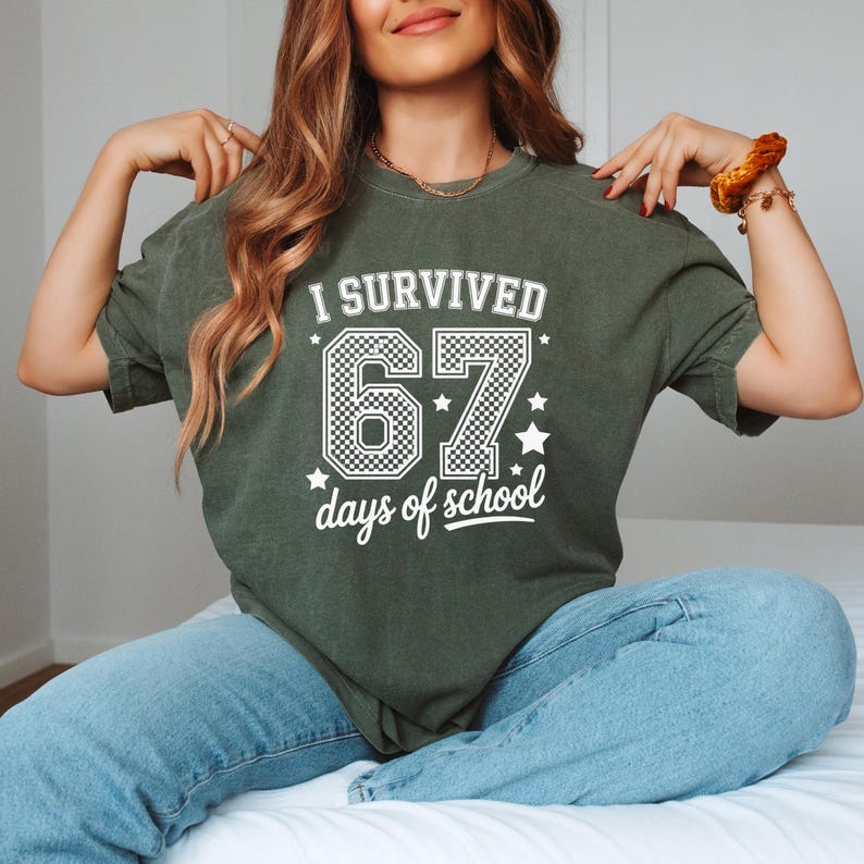 May include: A dark green t-shirt with the text "I Survived 67 days of school" in white. The number 67 is in a checkered pattern. The shirt is a crew neck and has short sleeves.