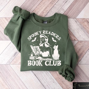 May include: A green sweatshirt with a white graphic design that says "Spooky Readers Book Club". The graphic features a woman reading a book with a cat sitting next to her.