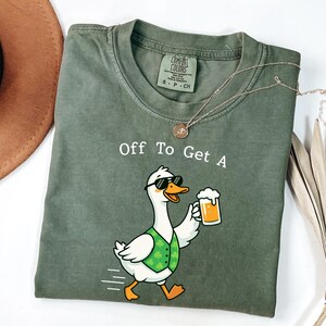 May include: Olive green t-shirt with the phrase "Off To Get A" and a cartoon goose wearing sunglasses and a green vest, holding a beer. A gold necklace is draped across the shirt. The shirt is a Comfort Colors brand.