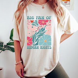 May include: Cream-colored t-shirt with a floral graphic and the text "BIG FAN OF HUMAN RIGHTS." The design features pink, blue, and red flowers with teal leaves. The shirt is a casual, comfortable style.