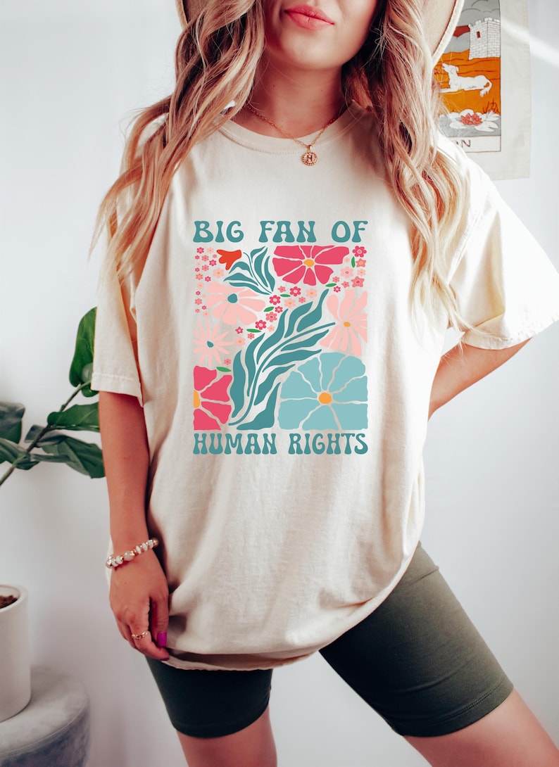 May include: Cream-colored t-shirt with a floral graphic and the text "BIG FAN OF HUMAN RIGHTS." The design features pink, blue, and red flowers with teal leaves. The shirt is a casual, comfortable style.