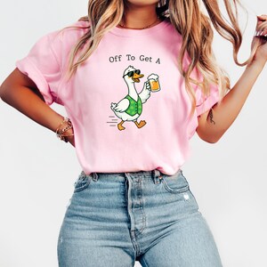 May include: Pink t-shirt with the text "Off To Get A" above a cartoon goose wearing sunglasses and a green vest, holding a beer. The goose is walking with motion lines.