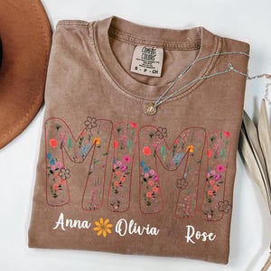Pode incluir: Camiseta marrom com a palavra "MAMA" em um design floral. Os nomes "Anna", "Olivia" e "Rose" s&atilde;o impressos abaixo. Um colar de prata com um pingente est&aacute; na camiseta.